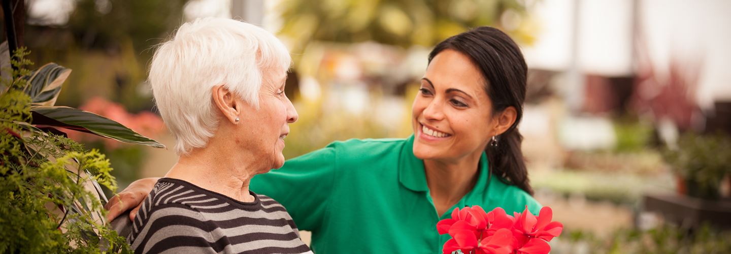 An elderly lady sitting outside on a bench with a Caregiver whilst holding some flowers