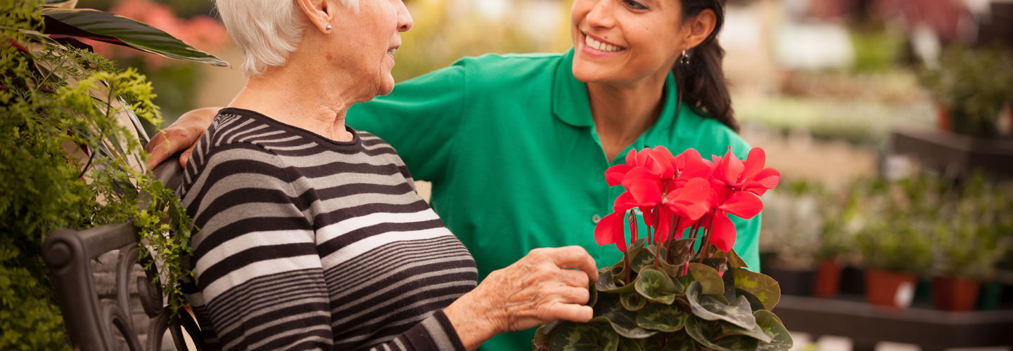 An elderly lady sitting outside on a bench with a Caregiver whilst holding some flowers
