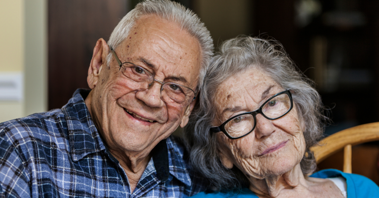 Elderly couple sitting together at home, with the woman leaning her head gently on her partner in a moment of comfort and companionship.