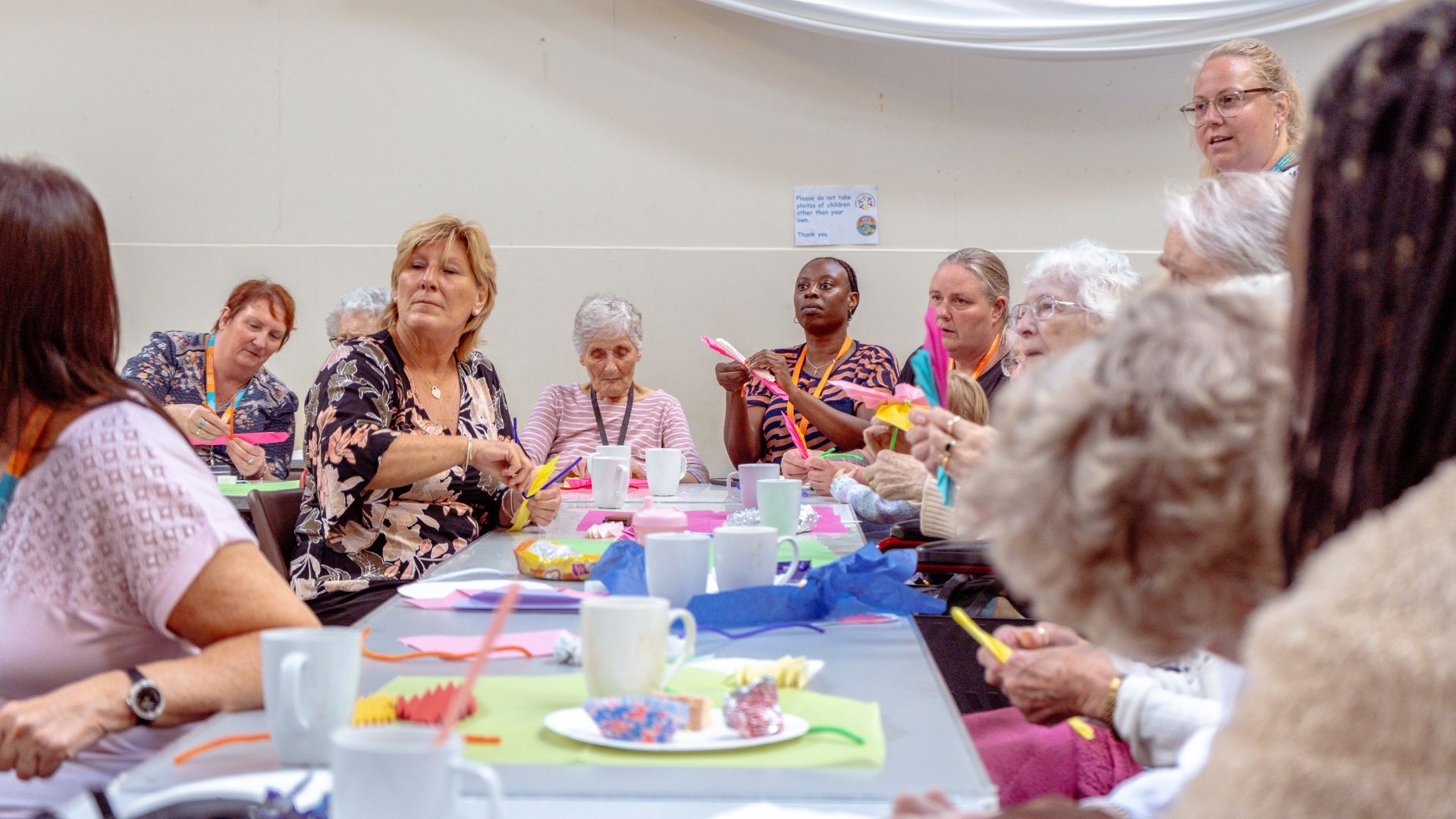 Care assistants and clients sitting at a table in Rowner Parish Hall, Gosport, making paper butterflies during the First Summer Homecare Coffee Club to promote social interaction and creative activities.