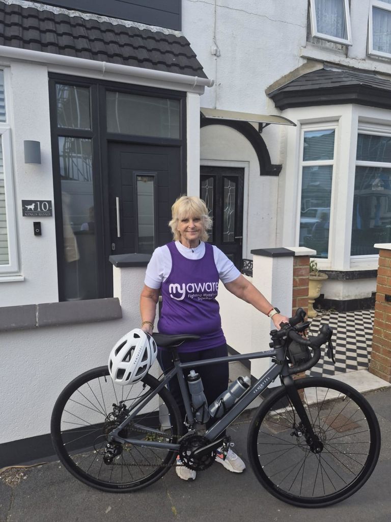 Julie standing beside her bicycle, smiling and holding the handlebars during her charity ride preparation for Myasthenia Gravis awareness