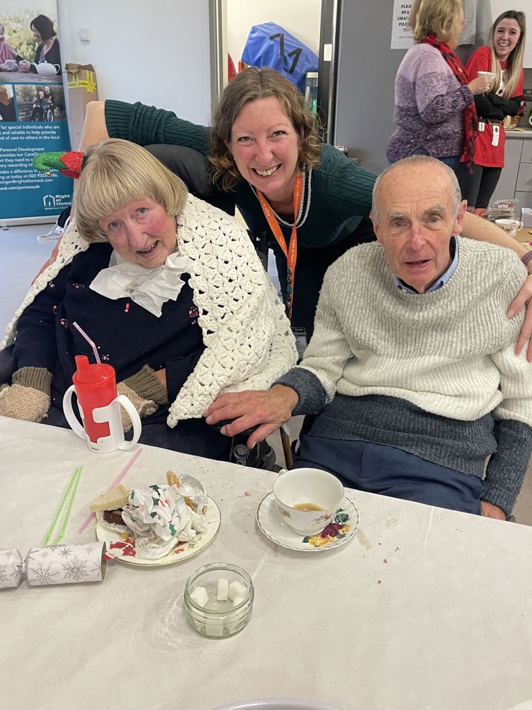 Carer with Right at Home Portsmouth Clients who are husband and wife,  at a Client Afternoon Tea Event that is run annually. The carer is smiling and has her arms around the couple as they sit on the table after eating cake and drinking tea.