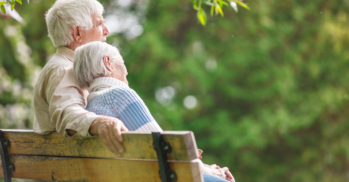 Elderly couple sitting outdoors in a garden with trees, the woman leaning her head gently on her partner, showing comfort and companionship.