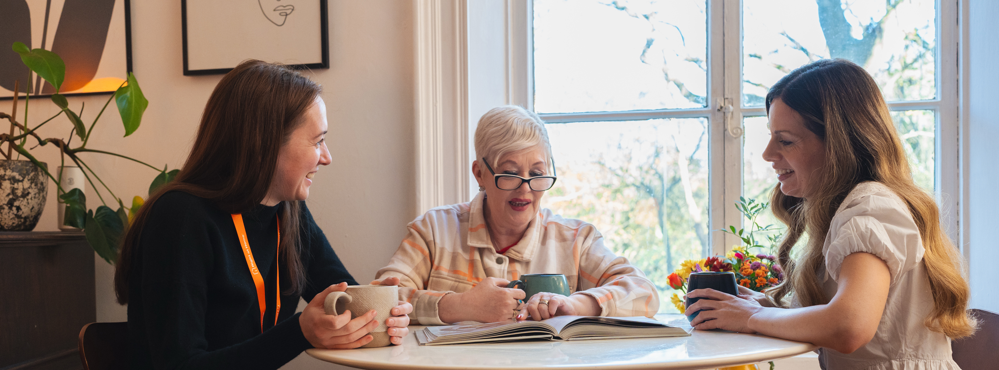 Caregiver and adult daughter sitting with elderly mother at kitchen table sharing tea and conversation at home