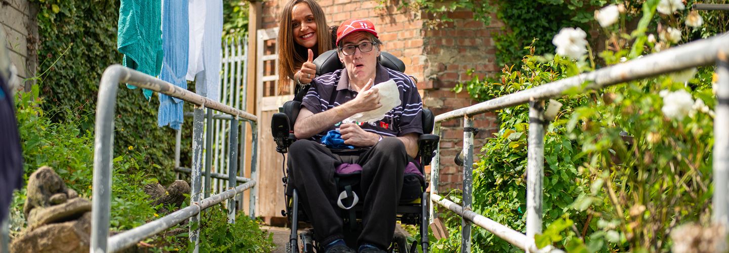 Client in wheel chair being pushed by a CareGiver through a garden