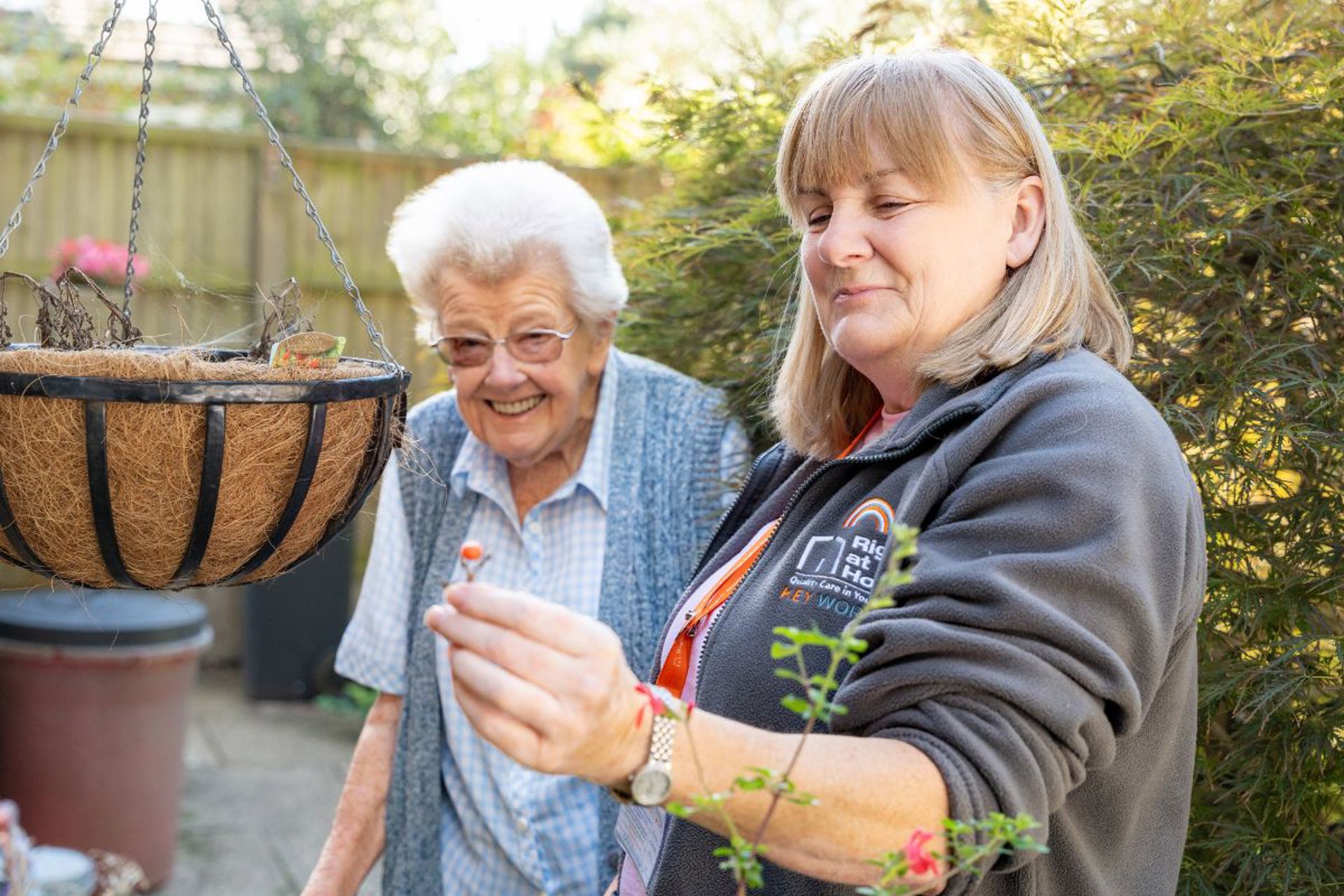 Carer showing a small flower to a smiling client outside their home, sharing a warm and friendly moment.